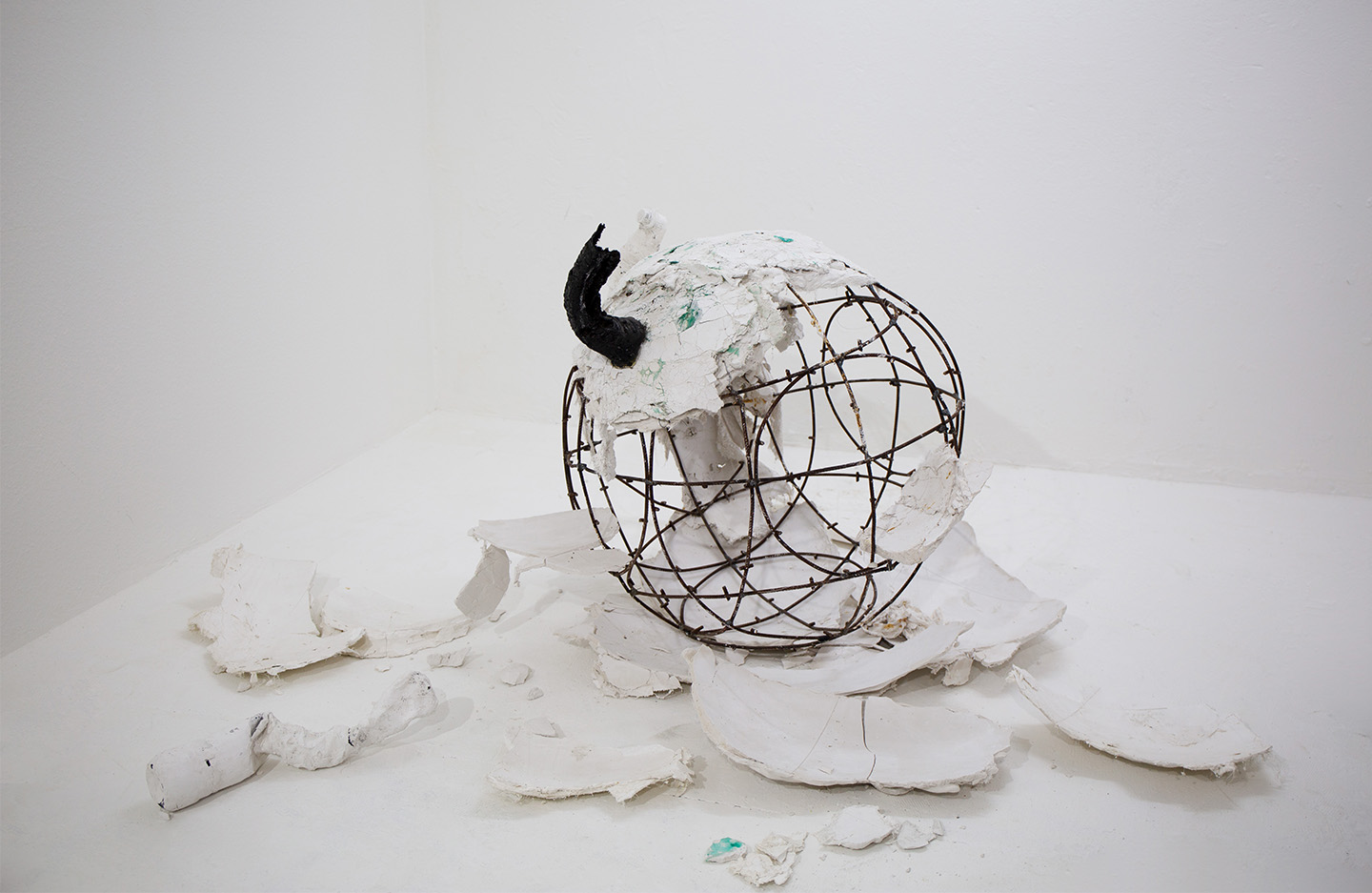 This image shows a sculpture  by Selou Sowe. The sculpture has a basic framework of steel struts welded together in rings to form a sphere. The framework is covered with what appears to be broken fragments of a former white sphere. There are also such fragments scattered on the floor. On top of the sculpture is an object that is small in relation to the sculpture. It is shiny black and inorganic.